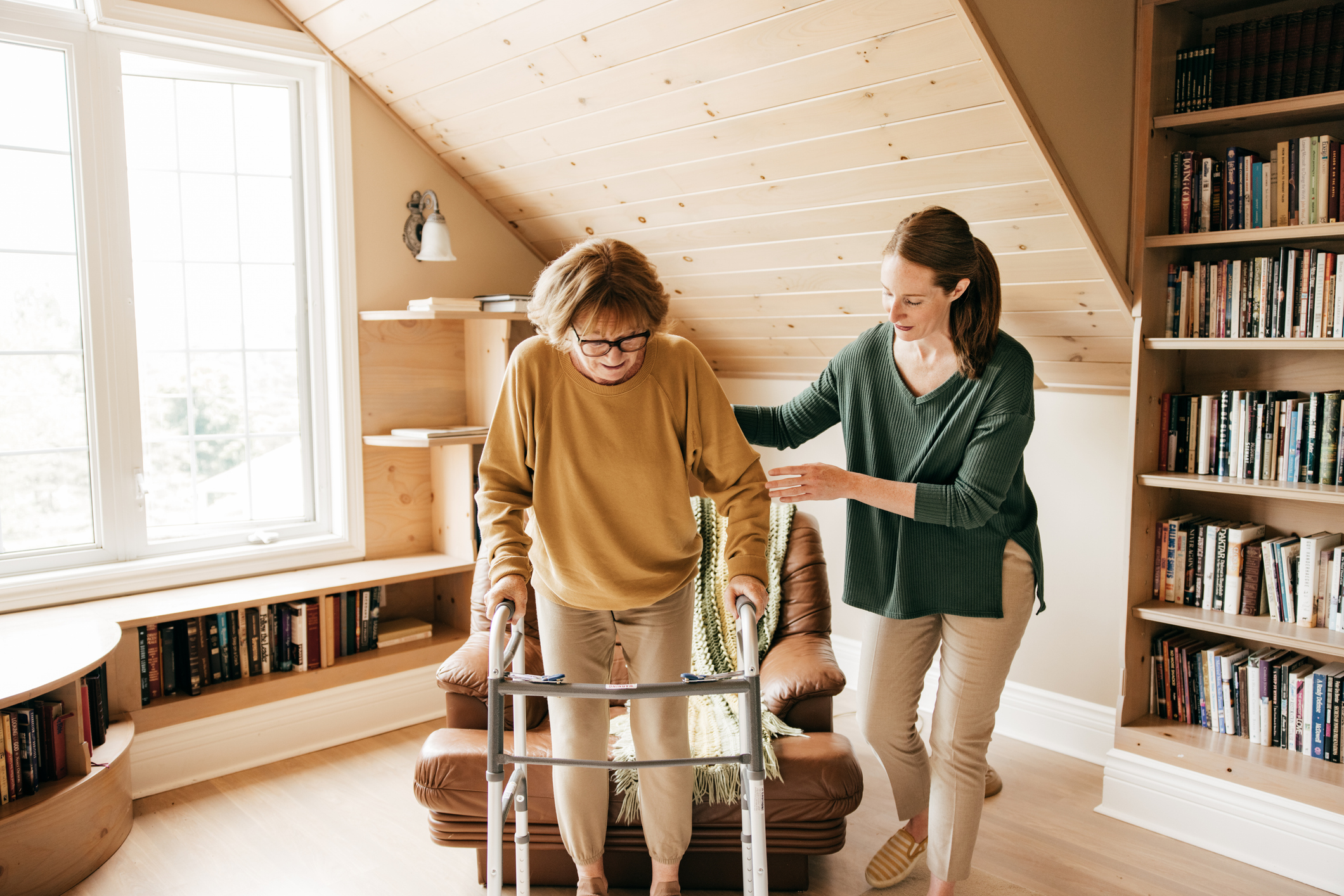 Young female caregiver assisting senior loved one safely transition from seated position to walker at home