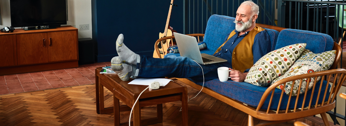 Senior man sitting on a couch in a music studio with laptop and coffee in hand
