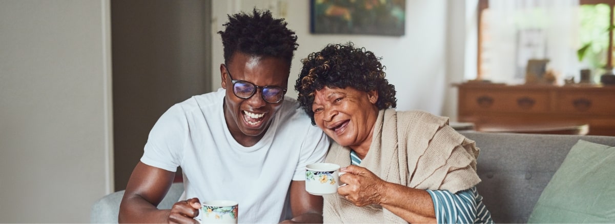 Senior woman and younger adult male sitting on the couch drinking coffee and laughing.