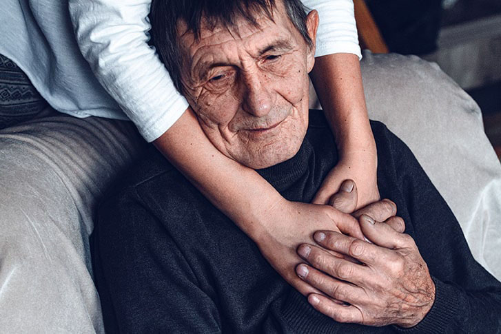 aging-man-in-chair-with-caregivers-arms-around-him-from-behind-SM-min.jpg