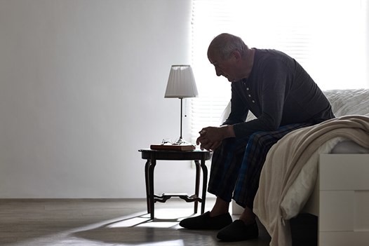 senior man sitting on unmade bed hands clasped head down