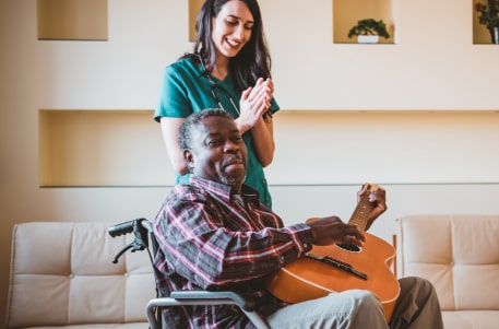 senior man in wheelchair playing the guitar while SYNERGY caretaker claps