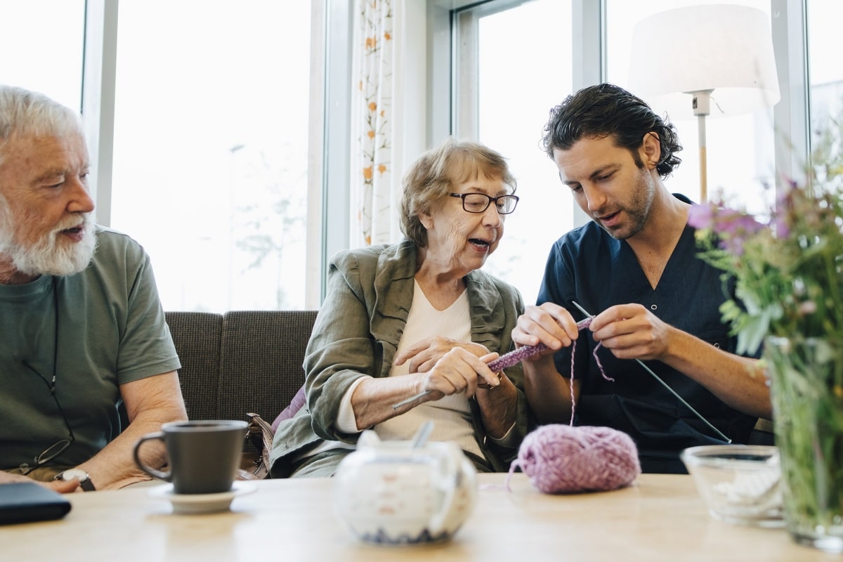Retired senior woman teaching knitting to male nurse while sitting on sofa at elderly care home