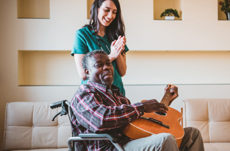 senior man in wheelchair playing the guitar while SYNERGY caretaker claps